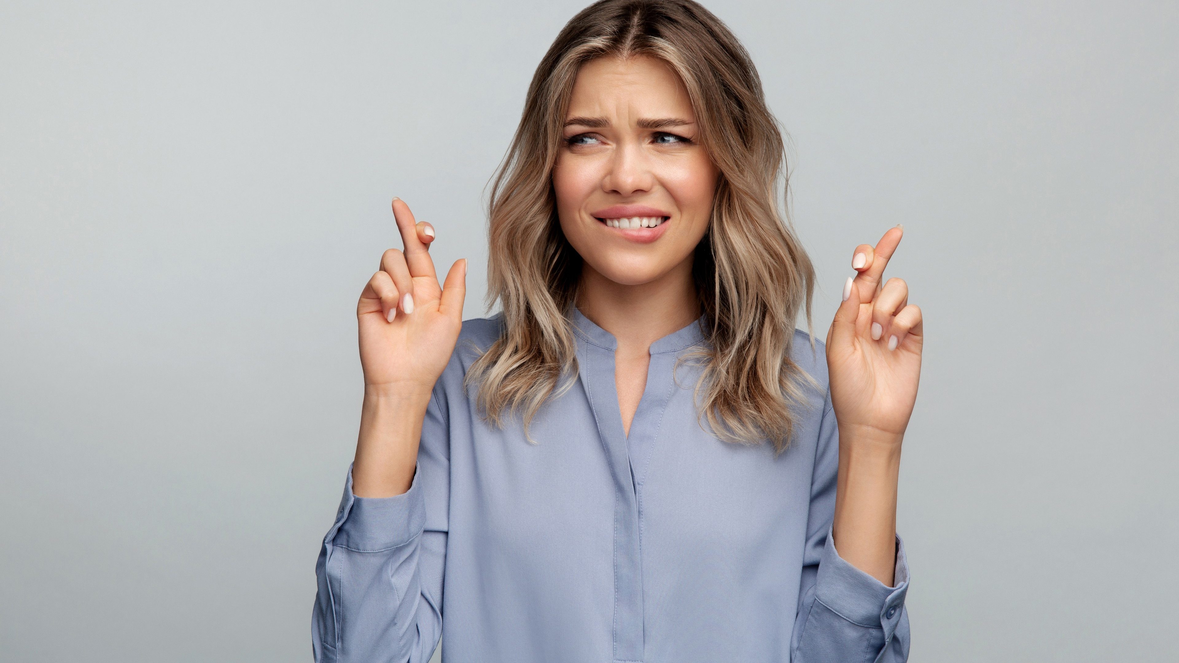 Stressful young woman crossing fingers and biting lip waiting for miracle, interview or exam result wishing to avoid troubles at work. Nervous female hoping for lucky solution isolated studio portrait