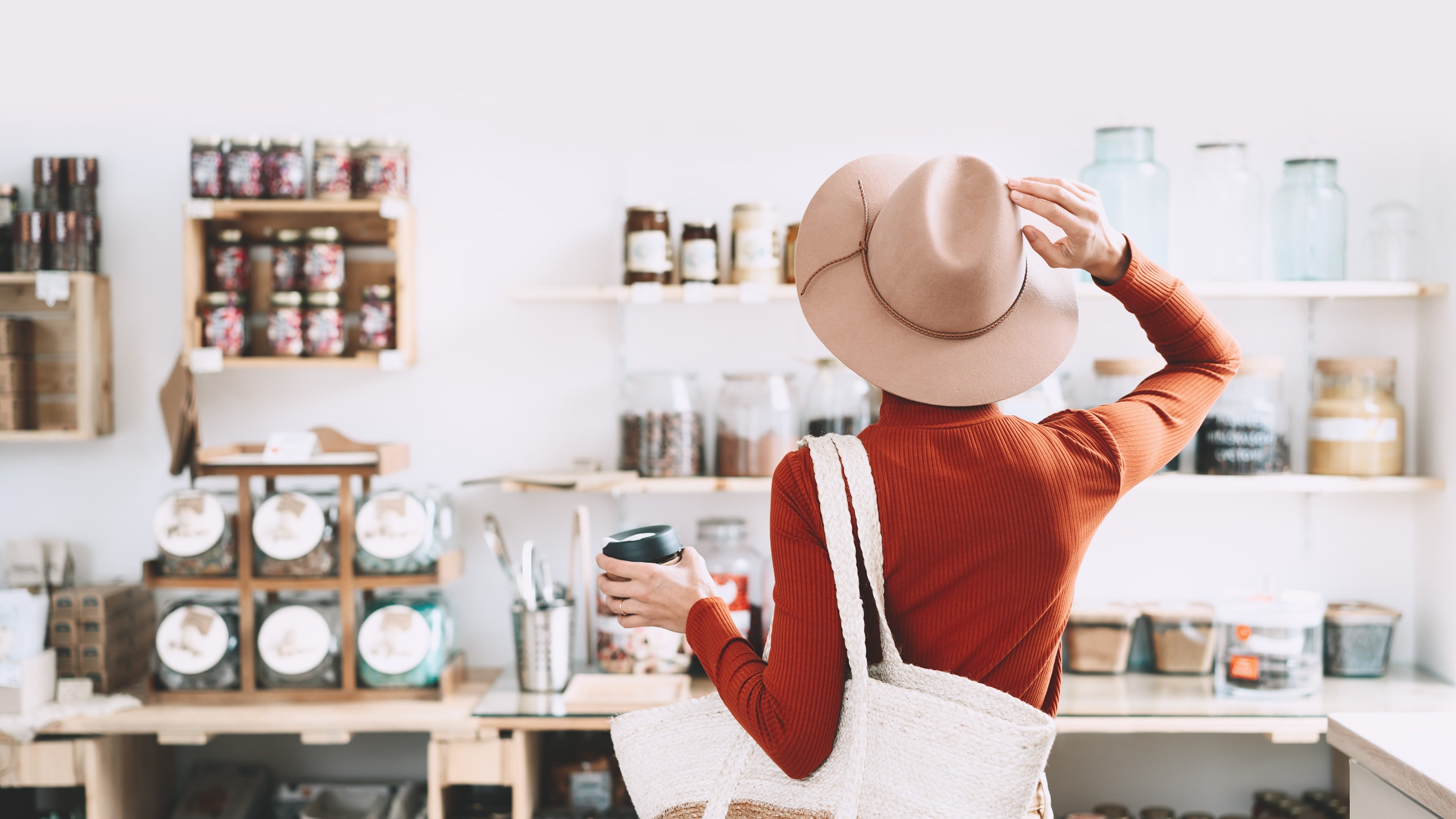 Minimalist vegan style girl with wicker bag and reusable glass coffee cup on background of interior of zero waste shop. Woman doing shopping without plastic packaging in plastic free grocery store.