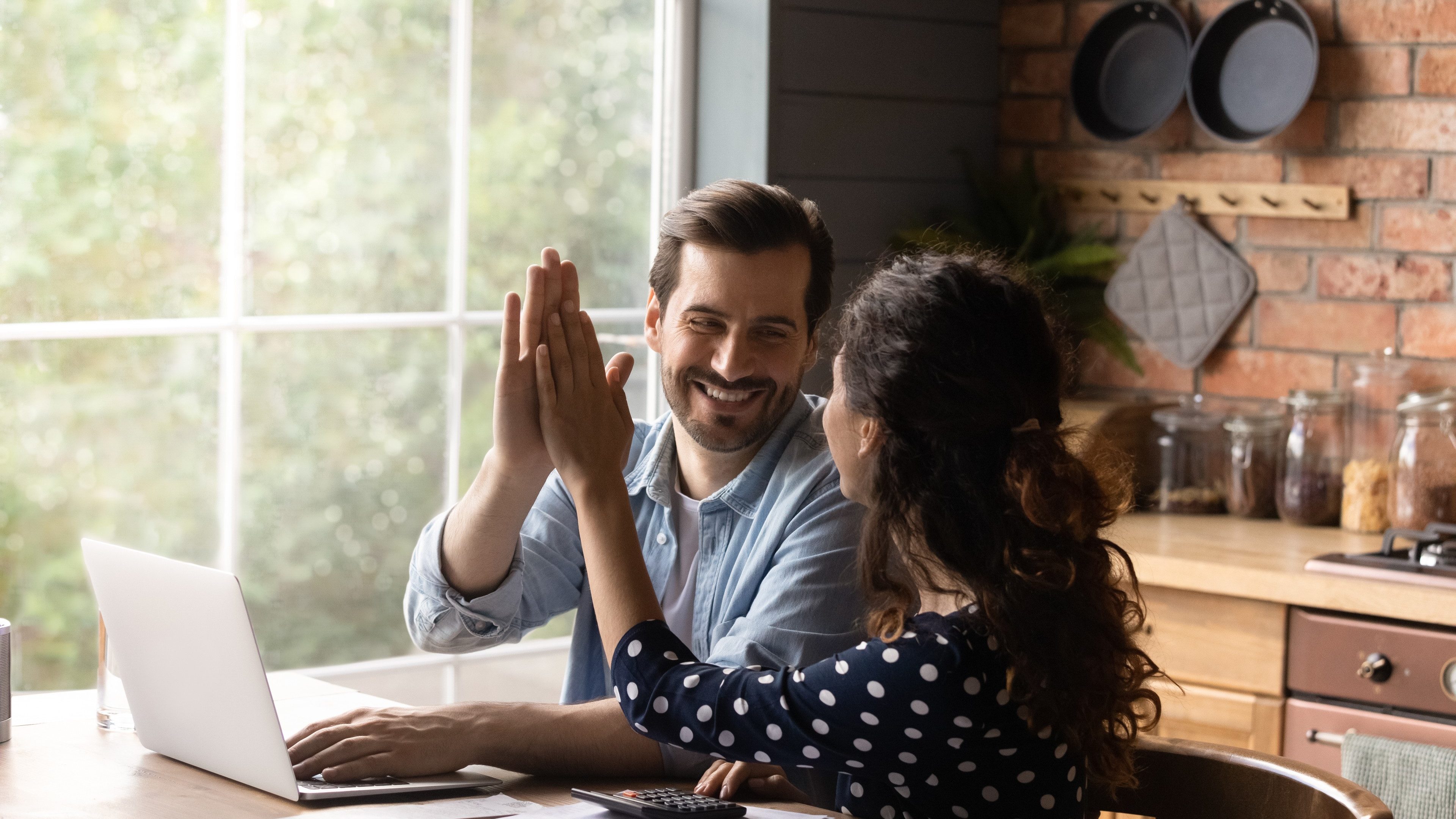 Happy young family couple giving high five to each other, finishing managing household budget, celebrating successful investment, making all payments online in e-banking computer application.