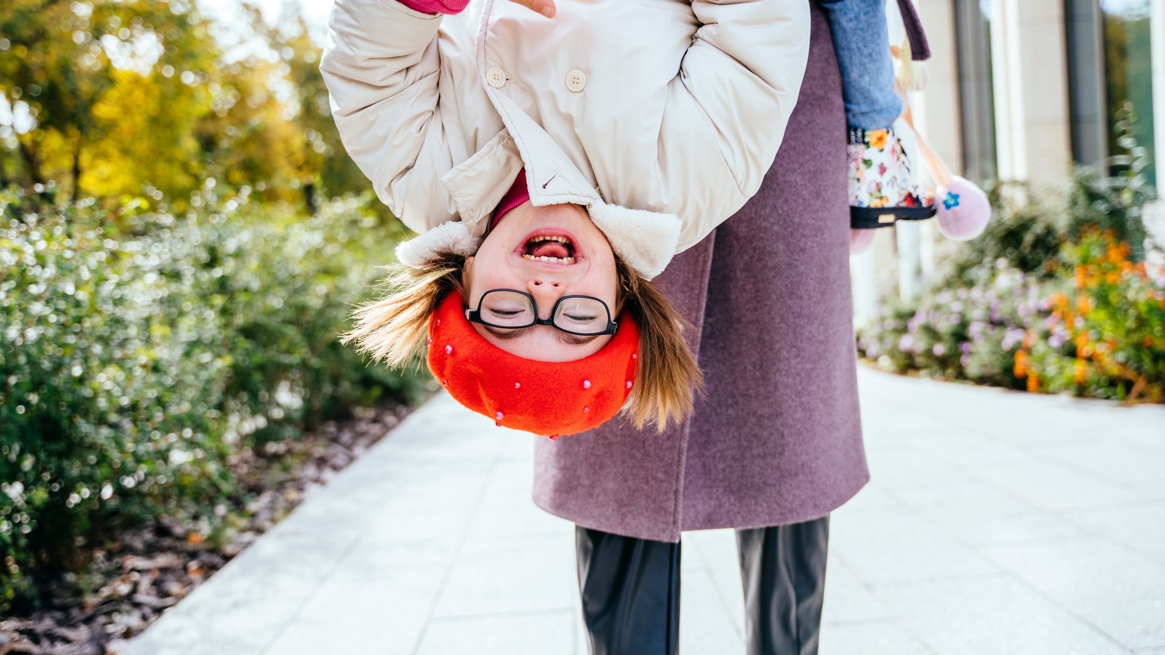 Brunette mom and cheerful adorable tot girl playing, having fun together in city street in autumntime, mother playfully holding her cute little daughter upside down.