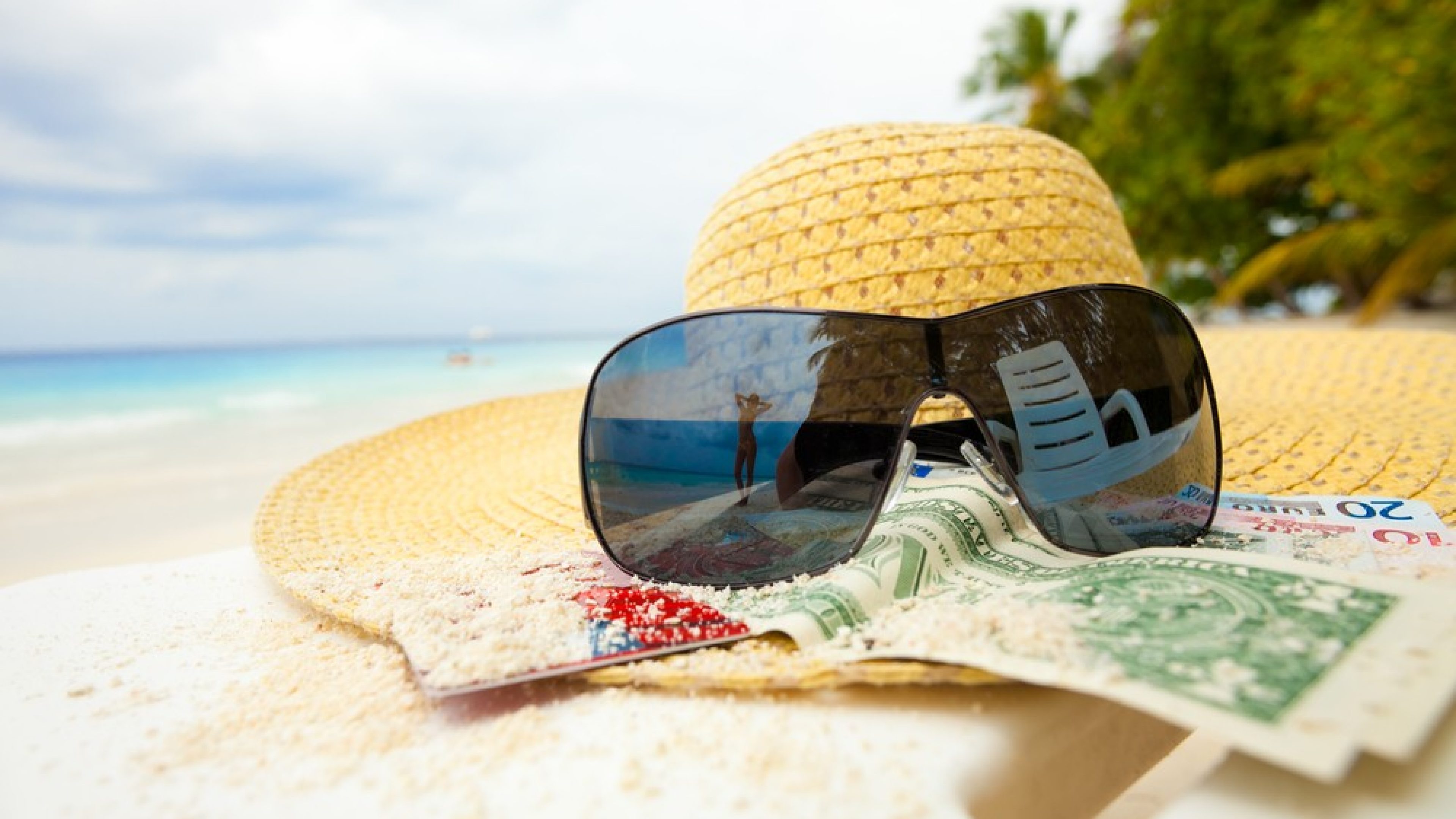 Straw hat, money, bank card, shades with relaxing woman mirrored in it
