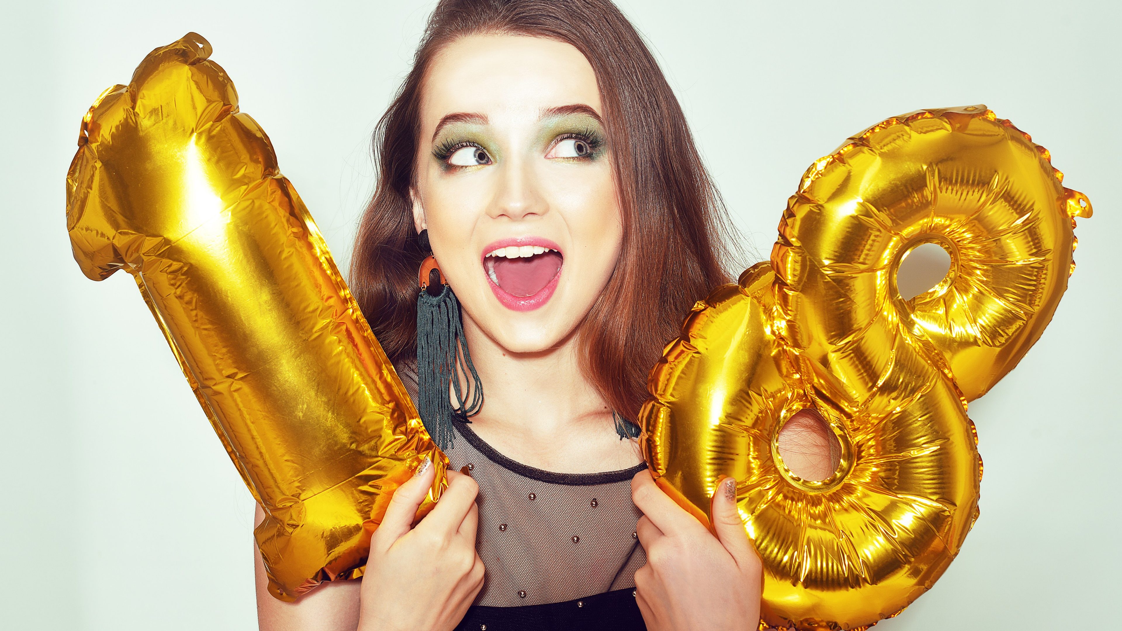 A birthday girl on her 18th birthday with gold number baloons. Excited eighteen girl with green make up and black dress smile. Model celebrating and having fun. Pretty cute girl on happy birthday.
