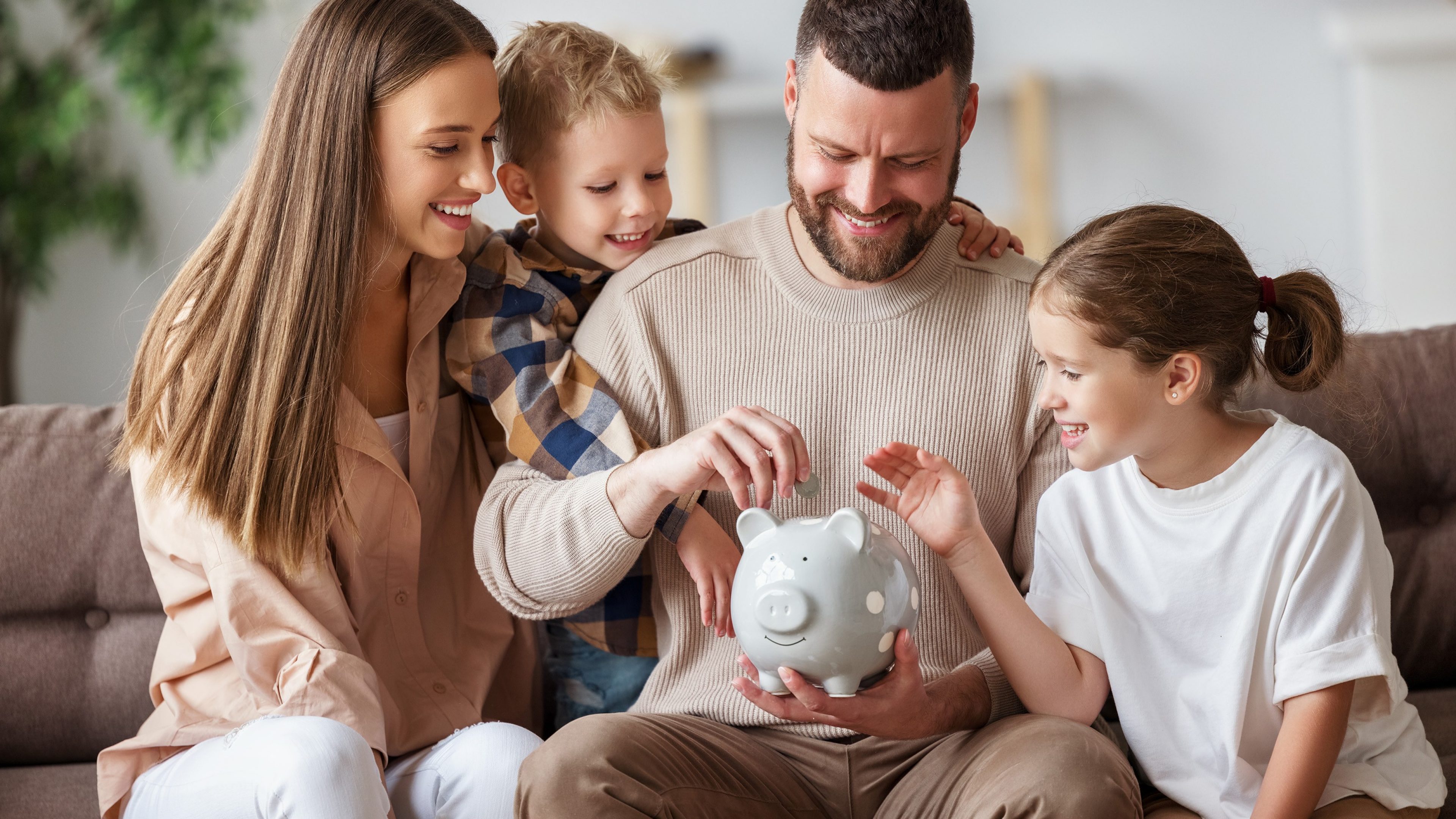 Happy family: cheerful mother and father with kids smiling and putting coins into piggy bank while sitting on sofa at home