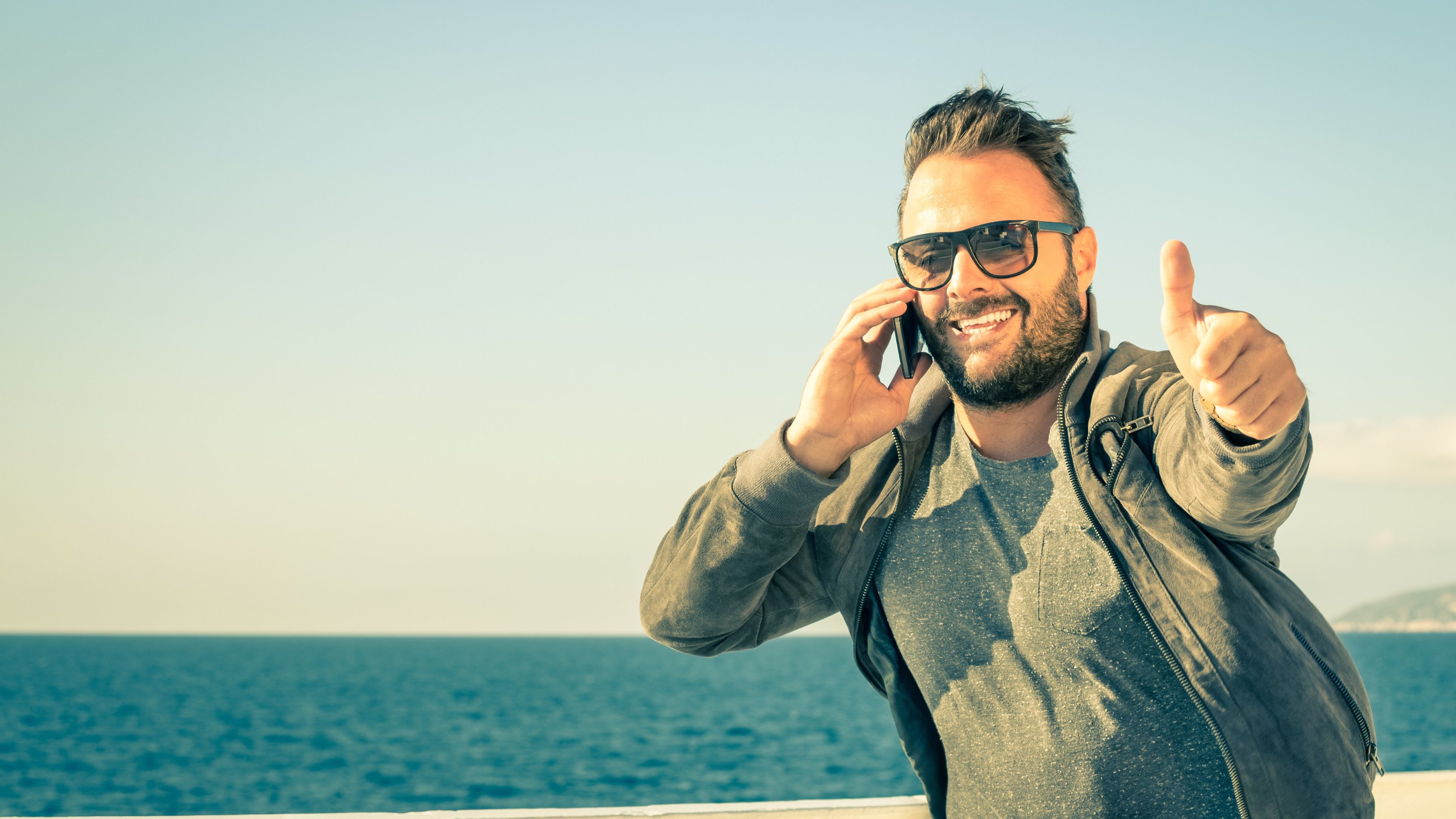 Young handsome man with thumbs up during a phone call with his smartphone - Concept of technology connected with traveller lifestyle - Male model showing success for a mobile telephony company