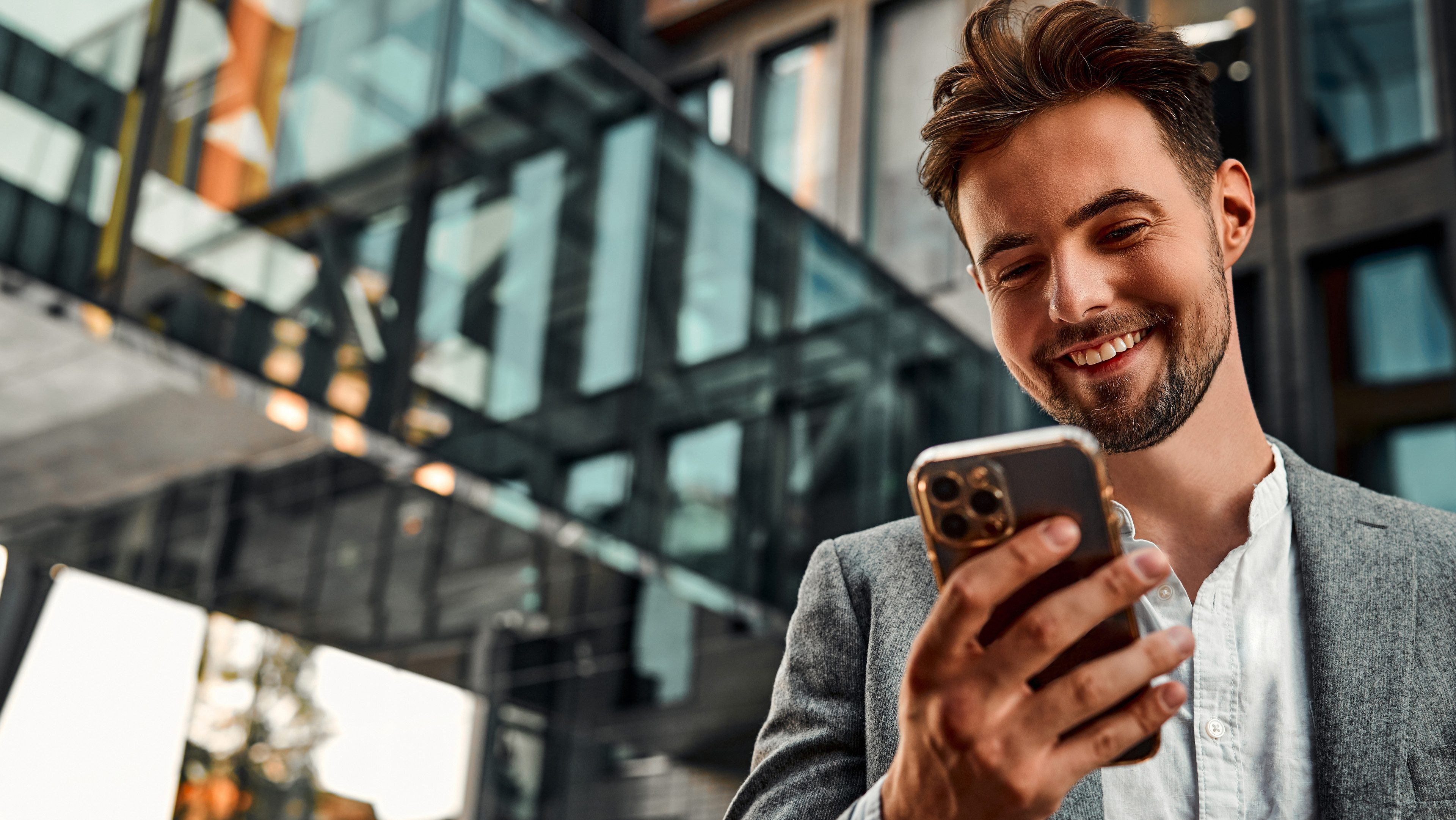 Portrait of attractive confident modern smiling happy adult dressed gray suit man holding a phone and standing outside near office building.  Copy space.                               