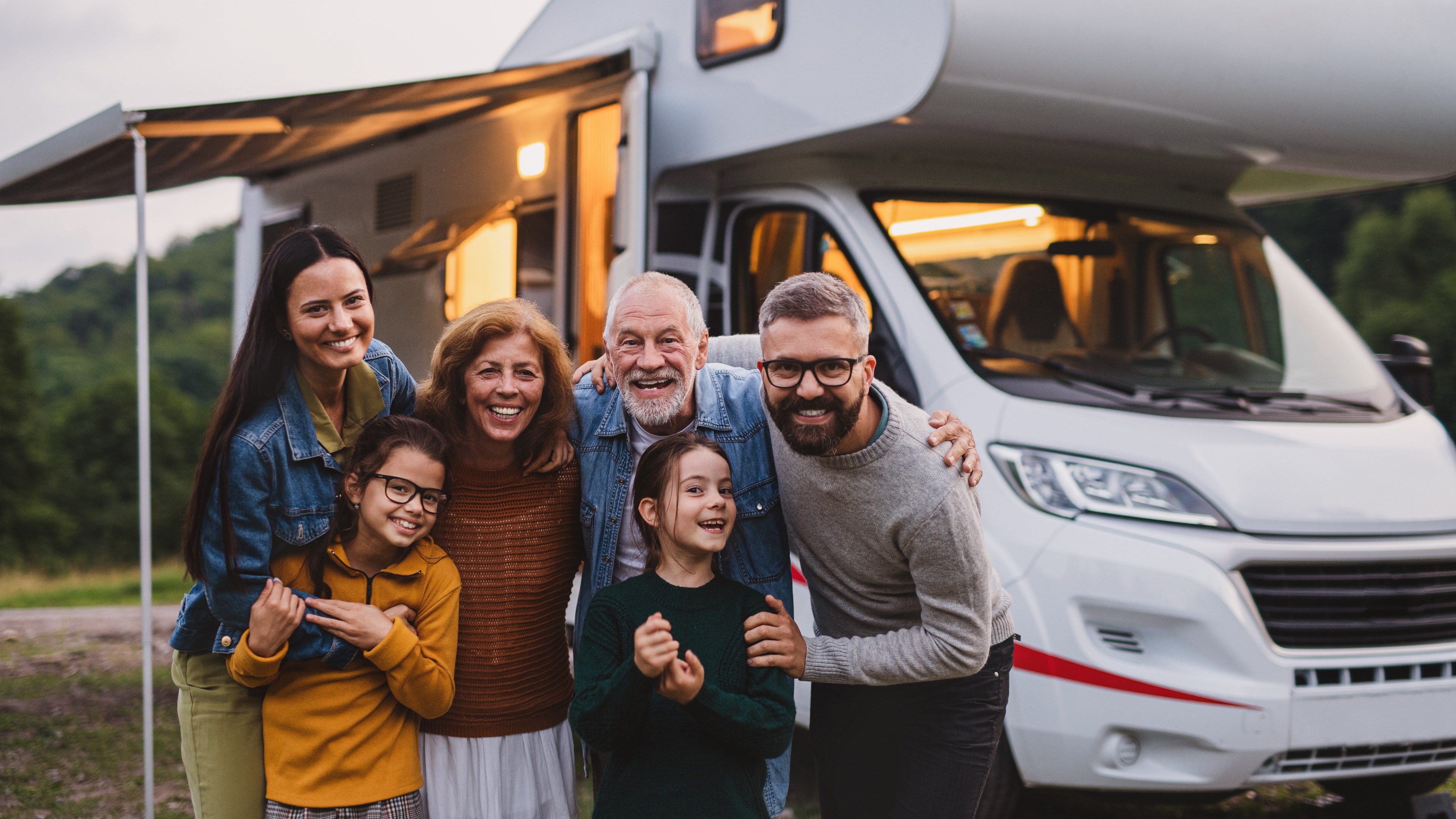 Multi-generation family looking at camera outdoors at dusk, caravan holiday trip.